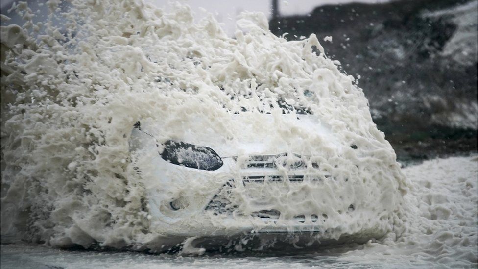 A car drives through sea foam whipped up by the wind of Storm Ophelia at Trearddur Bay, Holyhead