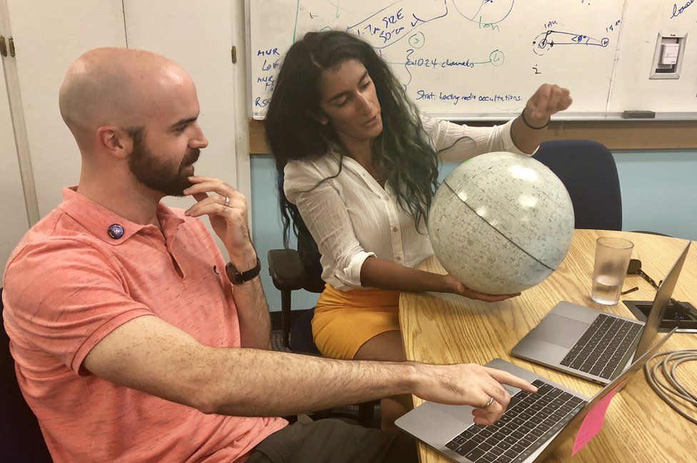 Two people look at a white globe of Mars. In the background there are whiteboards with calculations.