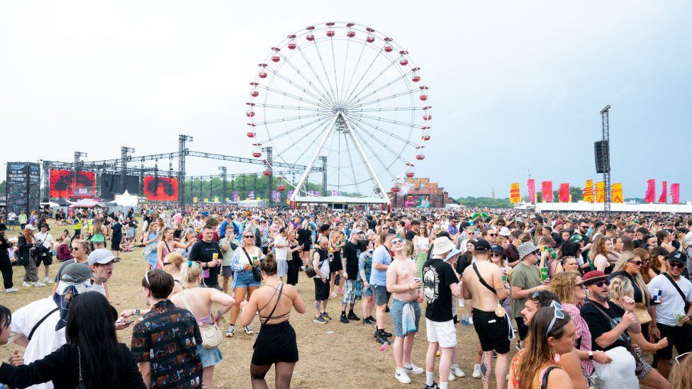 Сцена с фестиваля Parklife 2023. Большая плотная толпа людей заполняет большую часть переднего плана. В центре кадра над головой возвышается большое колесо обозрения. Слева металлические опоры сцены едва видны на фоне дымчато-голубого неба. Летом люди одеты в шорты, укороченные топы, а некоторые даже без рубашки.