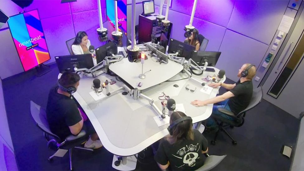 A picture of inside the Newsbeat studio with the presenter Pria Rai sat around a table alongside four young people who are seat around the table. Pria is wear a white jacket and has headphones on with a microphone and computer screen in front of her. The four other people are sat around the table from her and also have mics in front of them and headphones on.