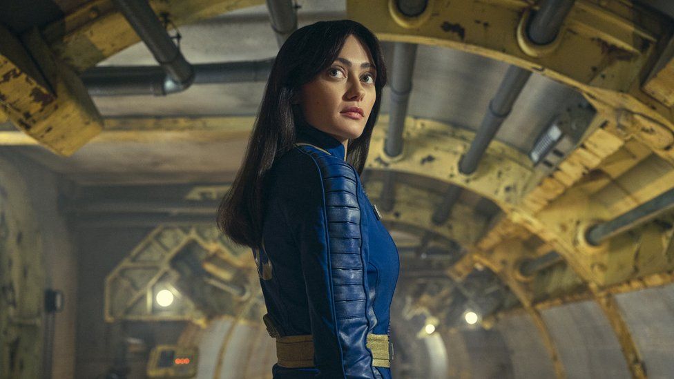 A young woman with long black hair stands in an artificially lit, cylindrical corridor with various pipes supported by bright yellow struts along its length She's looking back over her shoulder with an impassive expression. She wears a close-fitting, dark blue leather jumpsuit with panelling down the arms.
