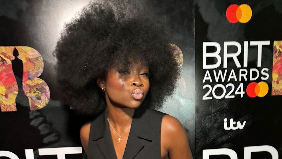 Baaba Nkansah-Asamoah. Baaba is a 20-year-old black woman with afro hair. She pouts at the camera, wearing a black waistcoat. She's pictured on the Brits red carpet in front of branded events hoarding
