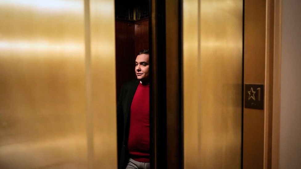 George Santos in red sweater between gold elevator doors.