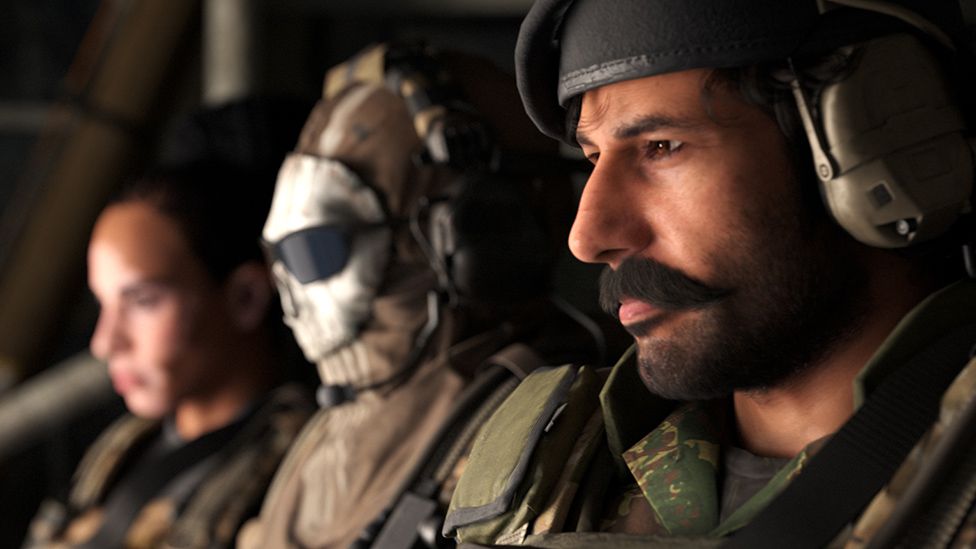 Three people sit, looking out of an aircraft or other military vehicle. Only the male nearest the viewer is in focus, the other two characters are blurred slightly. The man we can see clearly has a neat black moustache and light stubble on his chin. His brown eyes stare forward. We can just make out the earpiece of a pilot's helmet.