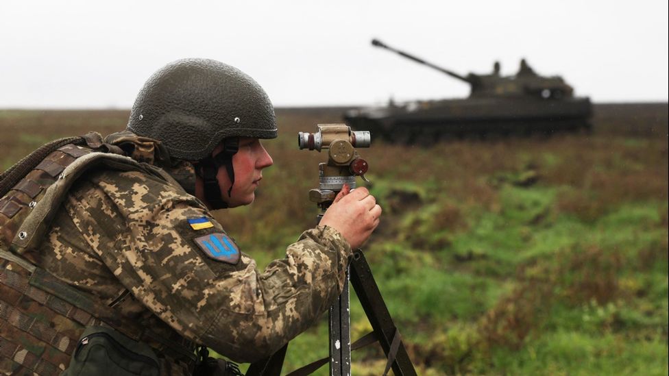 A Ukrainian soldier aims at a target with a self-propelled gun in the background