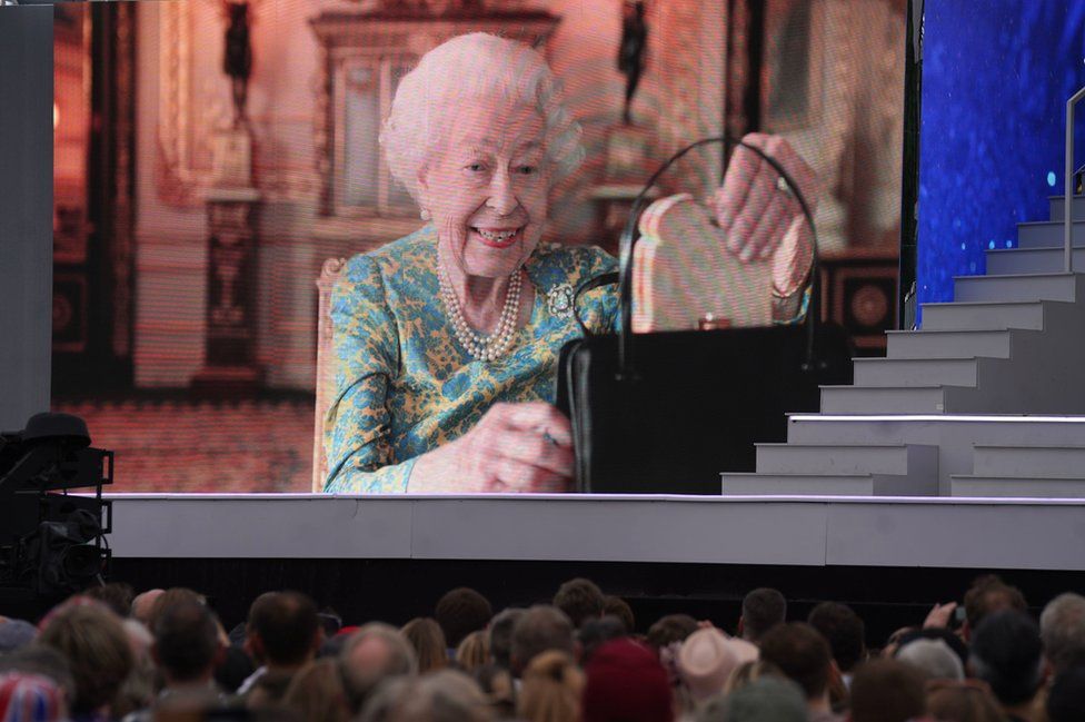The Queen producing a marmalade sandwich from her handbag, seen on a big screen outside Buckingham Palace