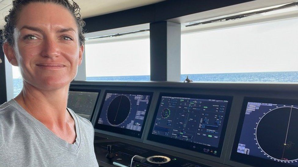 A white woman with tied back brunette hair wearing a grey t-shirt stands in front of the controls in the cabin of a superyacht. There are various screens displaying information about the weather and condition of the vessel. Through the windows behind her clear blue ocean can be seen.