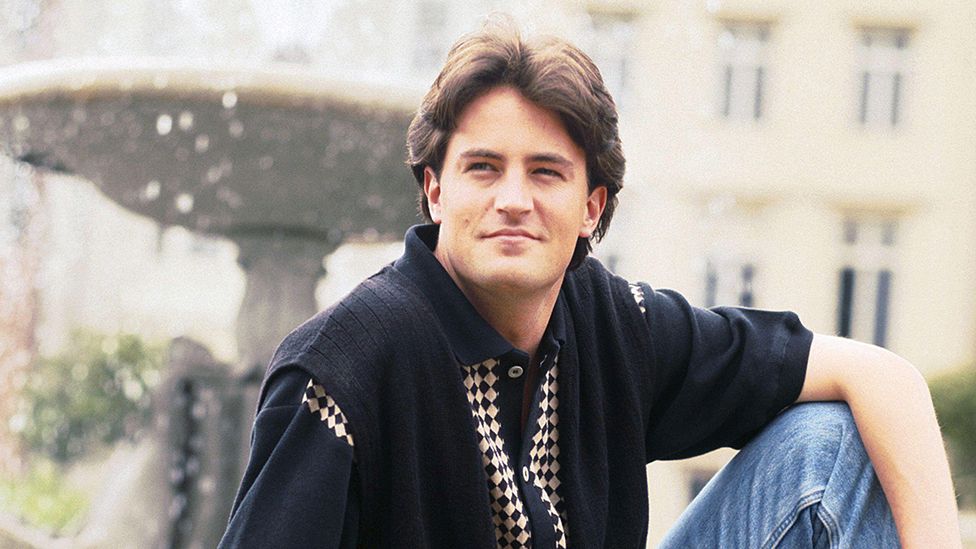 Matthew Perry poses for a portrait on a fountain, wearing blue jeans and a polo shirt