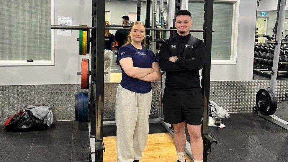 Phoebe Pothecary standing in front of a weight rack at the gym with her arms folded, beside another man