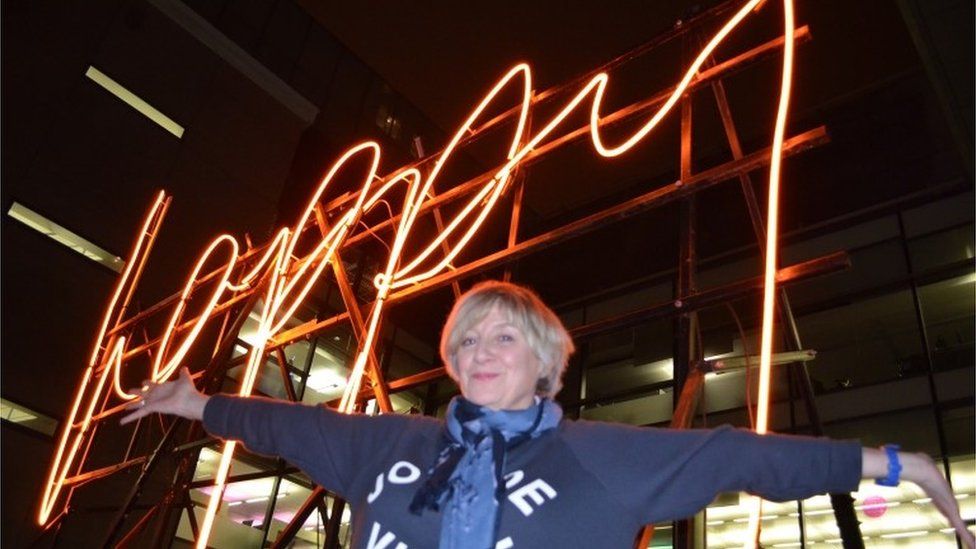 Victoria Wood in front of a neon sign saying 'happy'