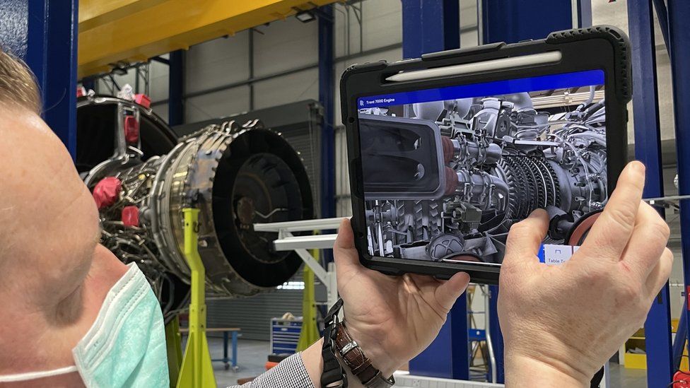 A tablet running augmented reality apps about a nearby engine is held by an employee