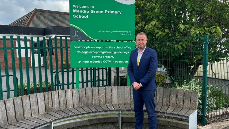 James Oakley wearing a blue suit standing by a school sign