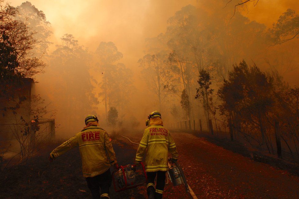 Two firefighters proceed along a road as heavy smoke fills the air, turning the sky a hazy orange