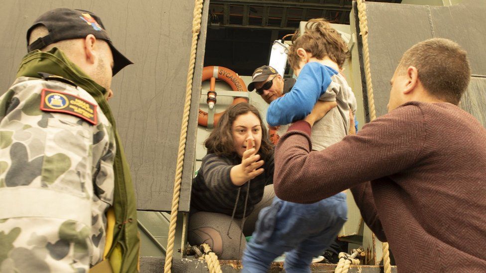 A member of the Australian navy looks on as a child is lifted on board a boat for evacuation from bushfires in Mallacoota, Victoria, 3 January 2020