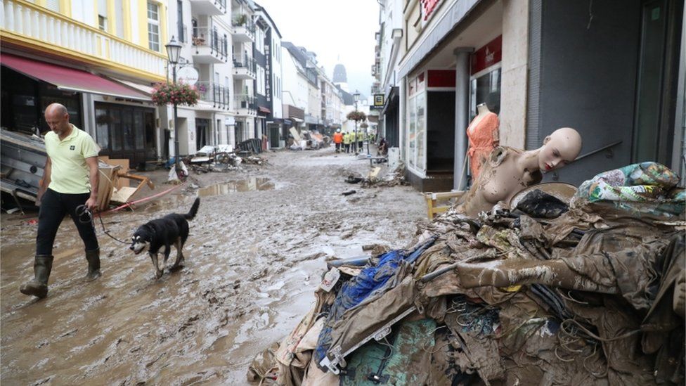 A person wearing rubber boots walks with a dog on a leash near debris sitting along a street after flooding in Bad Neuenahr-Ahrweiler, Germany, 16 July 2021
