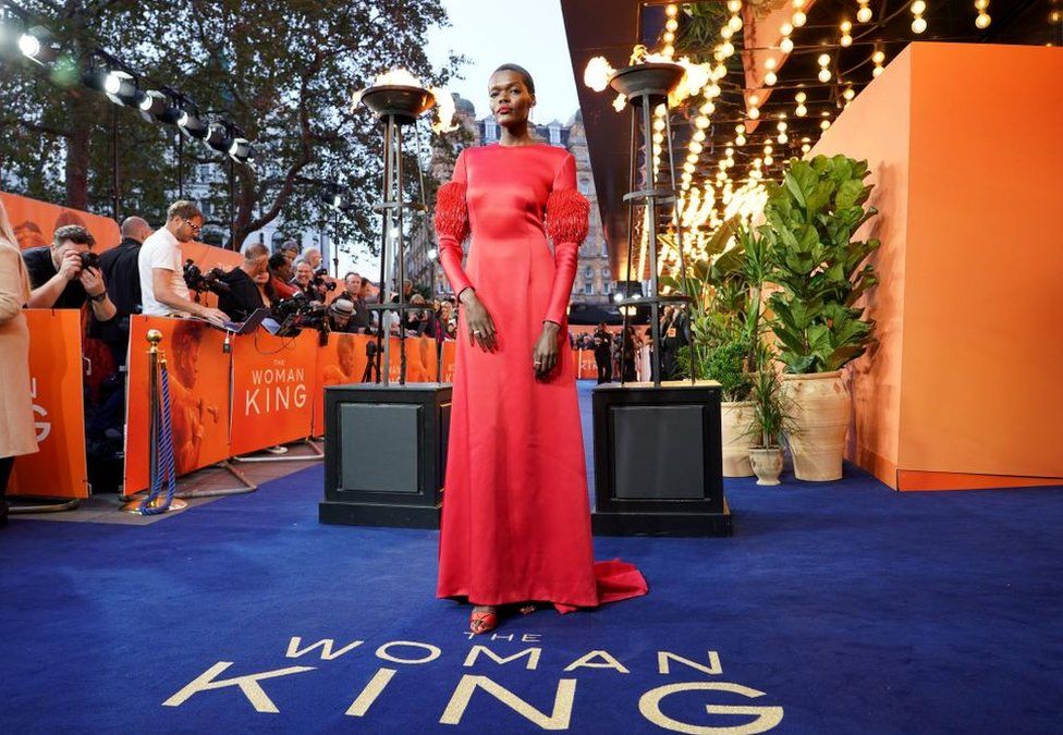Sheila Atim wearing a long red ball-gown dress staring into the camera. The blue carpet beneath her reads: The Woman King. There are lights behind her and photographers.
