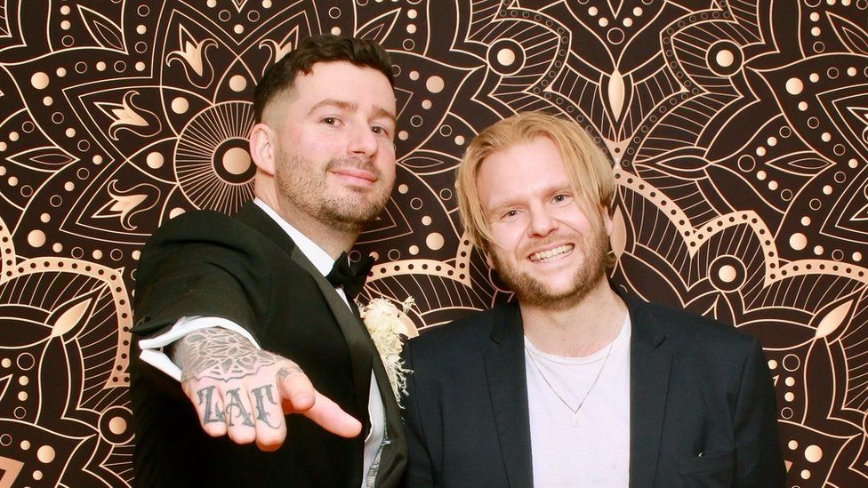 Tom Hollings and Jamie Osman. Tom, on the left, is a white man with short brown hair and stubble. Jamie, next to him, is white with longer blonde hair to his ears. They are both dressed in suits and Tom holds his tattooed right hand towards the camera. They are pictured in front of a patterned brown and gold backdrop