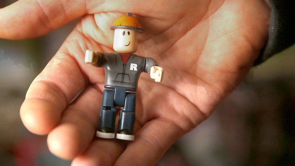 A hand holds a small model of plastic toy man with Roblox branding on it