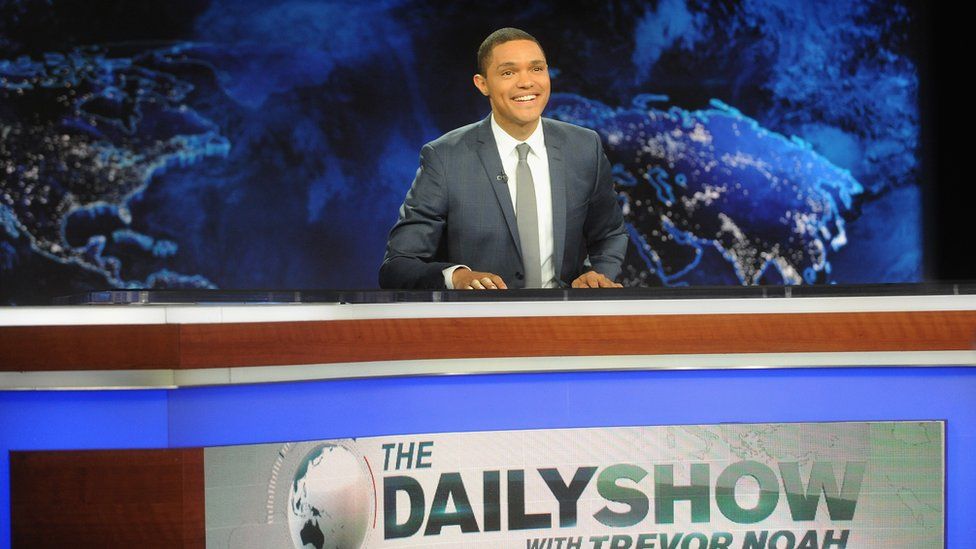 Comedian Trevor Noah behind a desk while hosting the Daily Show