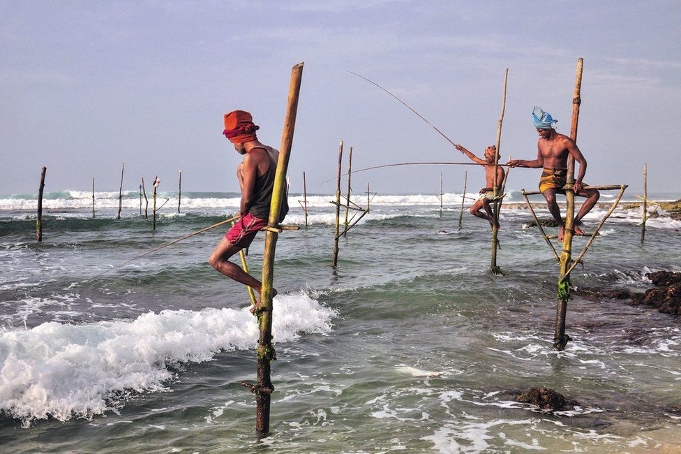 men perched on tall poles over waves on a beach, holding fishing lines