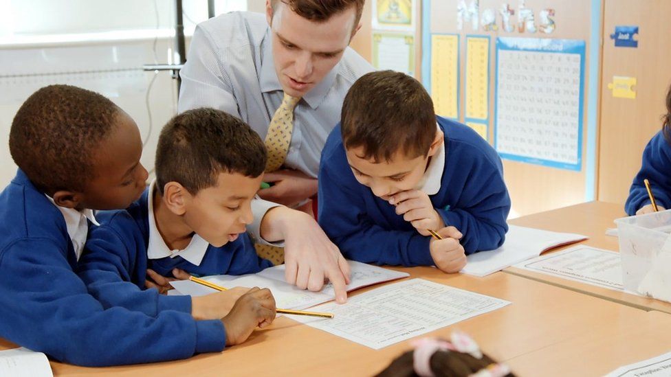 Man teaches three secondary school boys in classroom at their desk with work