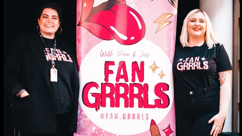 Jess Dyer (L) and Dan Hewitt (R) standing by a pull up pink and white banner saying Fan Grrrls on it