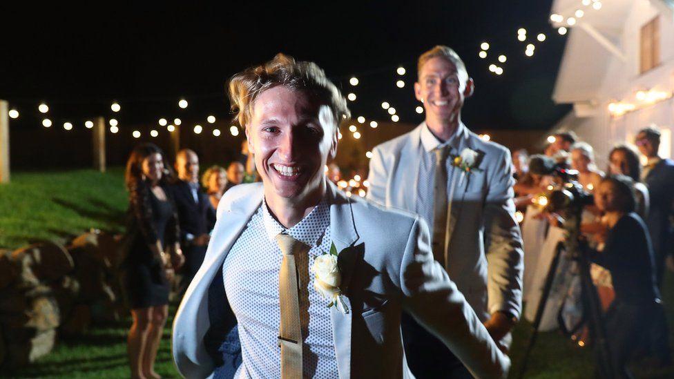 A smiling Luke Sullivan in front of his new husband at their wedding