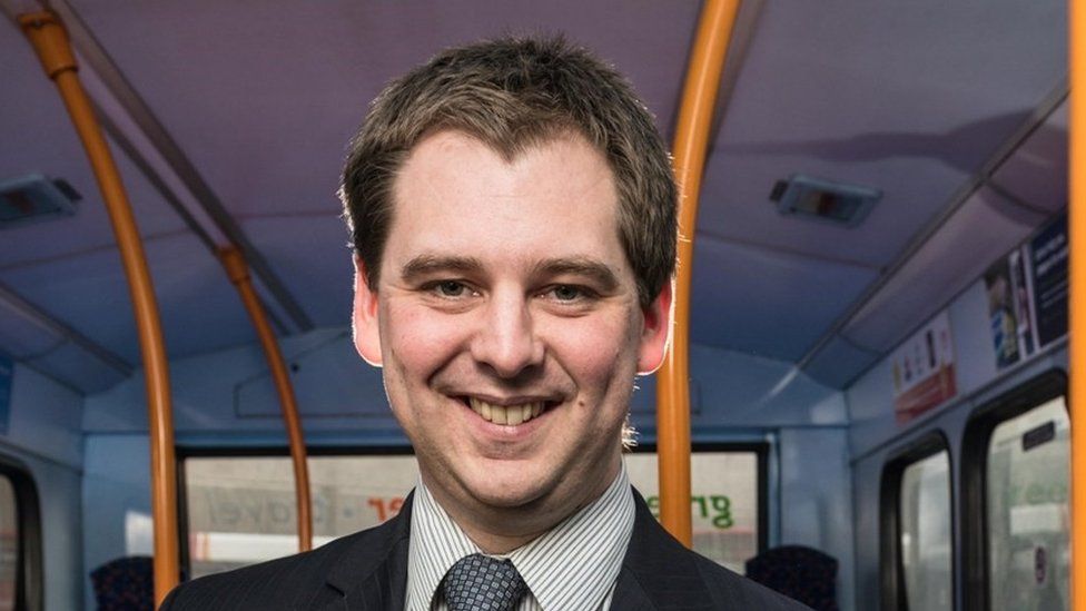 Mark Whitelocks with brown hair wearing a tie and standing in a bus