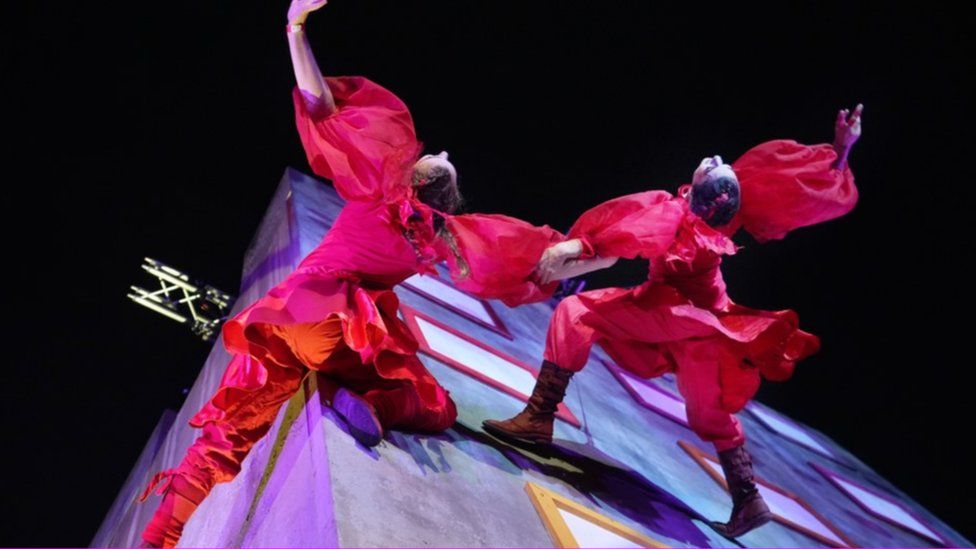 Two performers in red on wires walking vertically along the corner of a structure and linking arms with their heads thrown back
