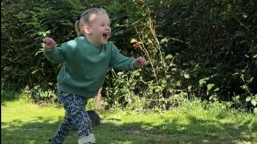 Young girl with ponytail wearing green top and blue trousers runs happily through grass.