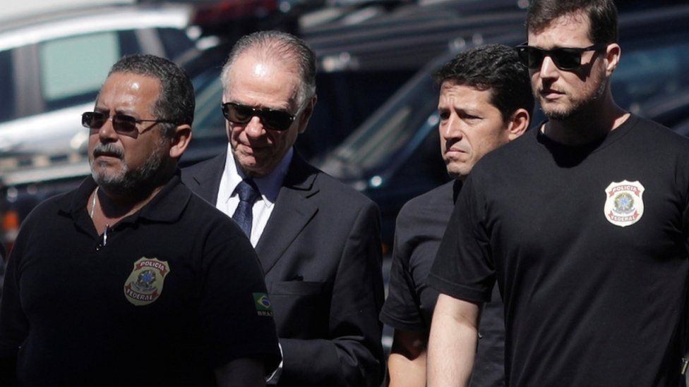 The President of the Brazilian Olympic Committee pictured between federal officers, wearing a suit and sunglasses