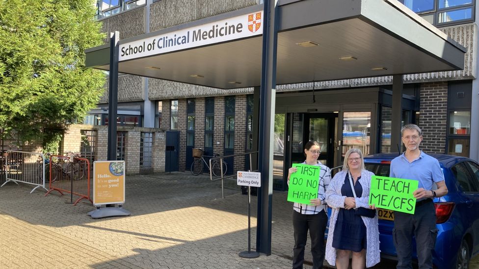 Three protestors holding green neon signs saying First Do No Harm and Teach ME/CFS in front of the School of Clinical Medicine in Cambridge