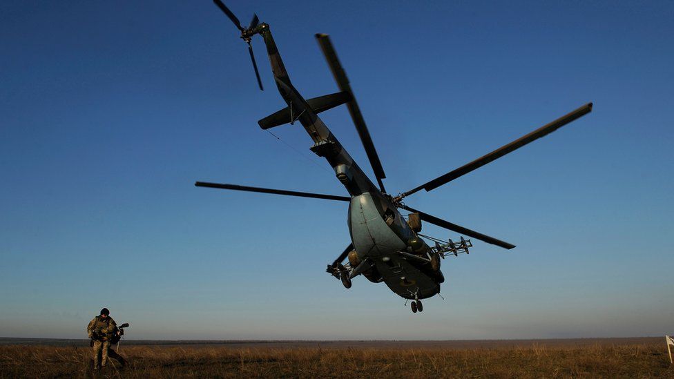 A serviceman walks near a Ukrainian military helicopter flying during military drills on an open green space with blue skies
