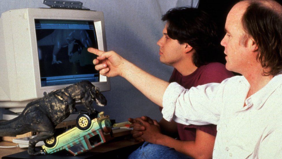 Two men look at a computer screen from the early 90s, with a computer animated dinosaur visible. A dinosaur model and jeep are on the desk beside them.