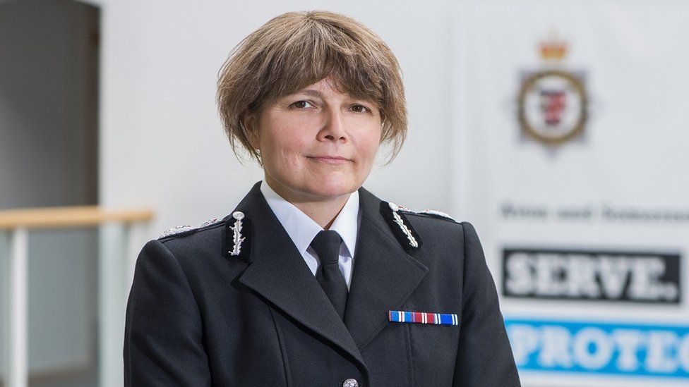 Avon and Somerset Police Chief Constable Sarah Crew. She is looking directly at the camera and smiling. Behind her, an Avon and Somerset logo can be seen, but it is out of focus. Ms Crew has brunette hair and is wearing a white shirt, a black tie and black suit jacket.