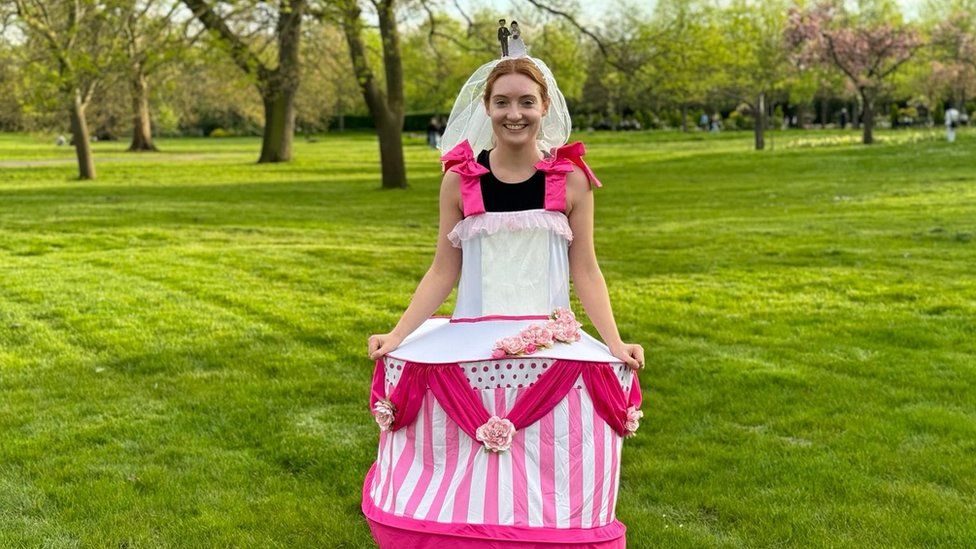 Laura Baker in her wedding cake fancy dress. Laura is a 27-year-old white woman with strawberry blonde hair which is tied back. She wears a bridal veil and has a wedding cake topper on her head. She has a black sleeveless running top with bright pink straps holding up her costume which is a white and pink tiered wedding cake. Laura's pictured outside in a park on a bright day