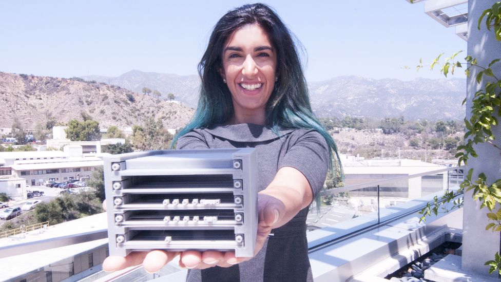 A woman with long, dark brown and green hair stands in front of a mountainous background holding out a grey, 3D printed model