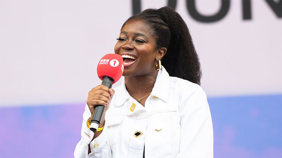 Clara Amfo, a black woman wearing a white jacket, holding a red Radio 1 microphone. The background is white and blue.