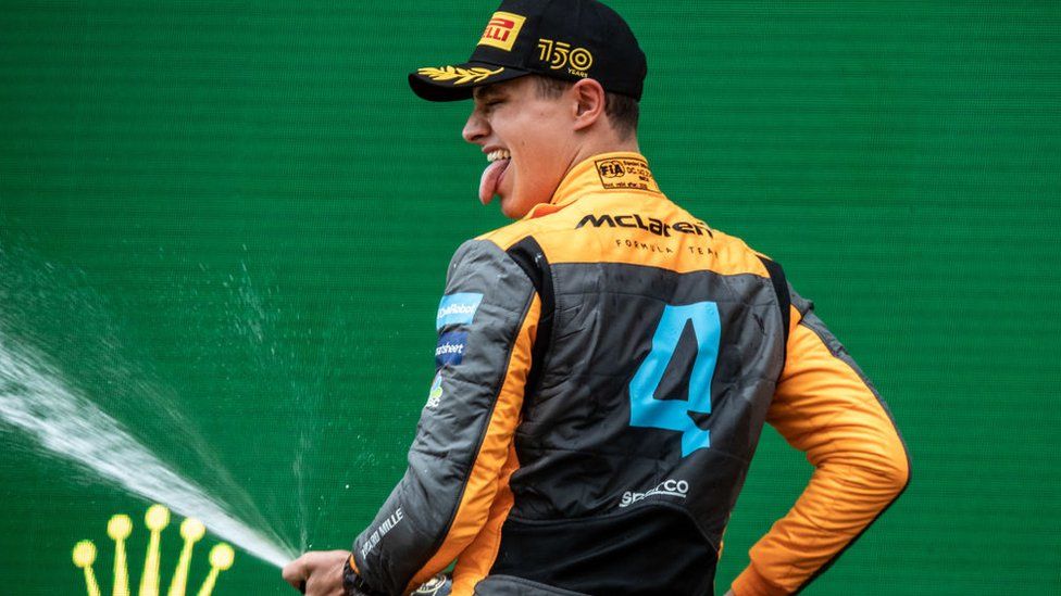 Lando Norris on the podium with his back to the camera. He is wearing an orange and black McLaren team jacket and a black and yellow Pirelli cap. He is sticking his tongue out and is spraying a bottle of champagne at waist height
