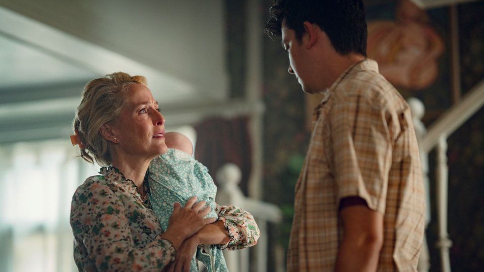 Gillian Anderson as Jean stands opposite Asa Butterfield as Otis. Jean, a blonde woman in her 40s, is carrying a baby dressed in a green patterned onesie over her shoulder. She has her hair clipped back and wears a floral blouse and looks up at her son with a concerned expression. Otis, a teenage boy with short dark hair, is visible from behind wearing a yellow plaid shirt. The characters are pictured in their home with a window and staircase visible in the background.