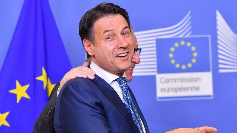 Italian prime minister Giuseppe Conte turns to camera, smiling, as he is led away by President of the European commission Jean Claude Junkcer