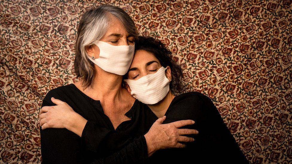 Two women embrace, wearing face masks