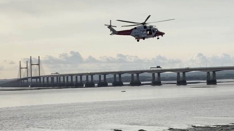 A helicopter hovering over the Severn Estuary with the Prince of Wales Bridge in the background
