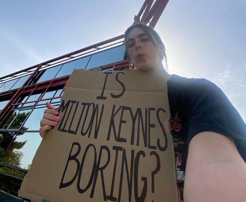 Anouska Lewis holding a sign that says "Is Milton Keynes Boring?" outside the Point, a former cinema