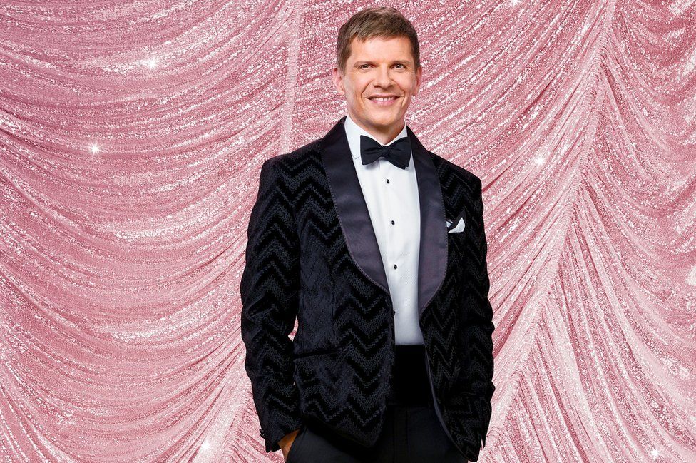 Nigel Harman in a black tuxedo with his hands in his pockets