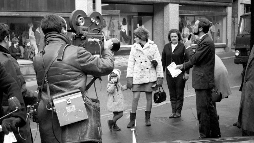 Man with beard holds microphone watched by shoppers and a film crew