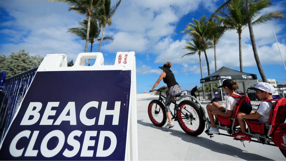 A cyclist with a trailer for children passes a "Beach Closed" sign on the boardwalk on March 22, 2020 in Miami Beach, Florida