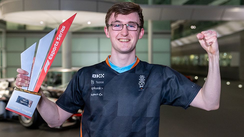 Lucas Blakeley stands, looking victorious, as he holds a triangular red and white trophy with "esports series" written vertically along it. He's smiling widely, with a large foyer in the background. The front end of a racing car is just visible in the background
