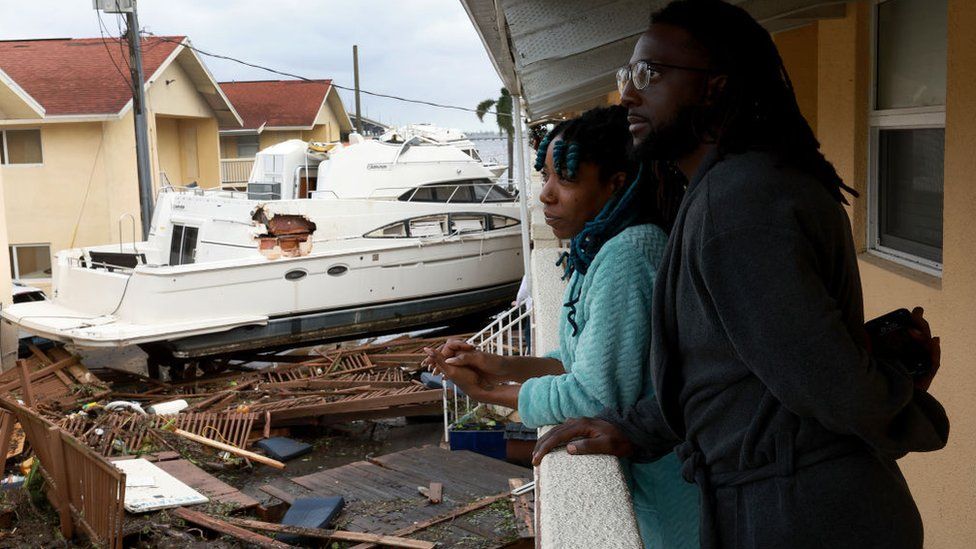 Мужчина и женщина стоят на балконе рядом с лодкой, которая врезалась в их жилой комплекс.