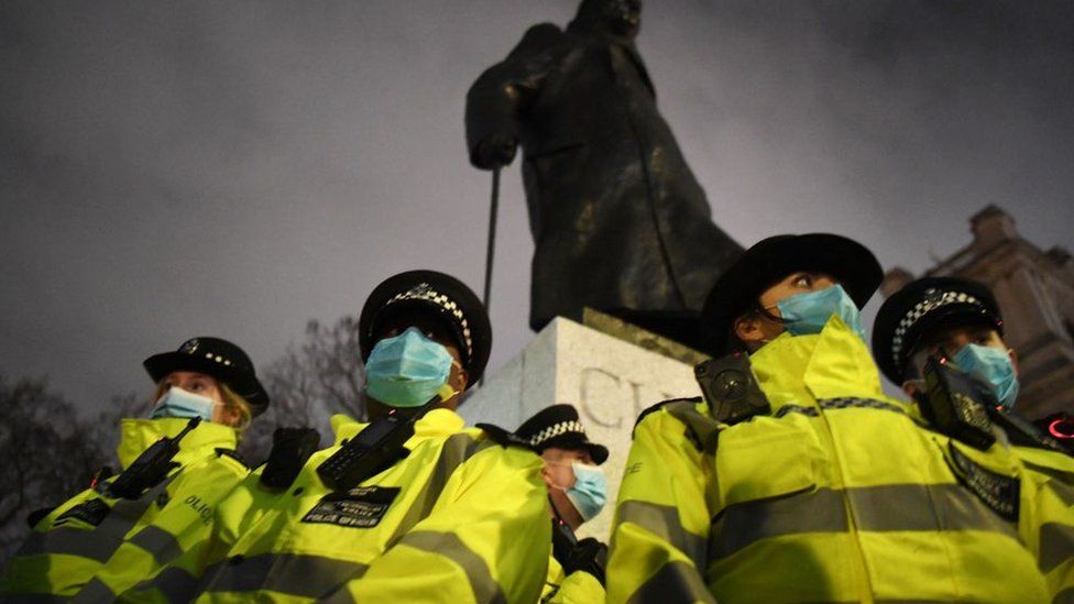 A group of Met Police officers dressed in reflective jackets and wearing Covid face masks stand in front of a statue of Winston Churchill with a cloudy night sky in the background